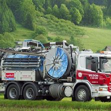Ein großer rot-weißer LKW mit 'WILTON' auf der Seite steht auf einem grünen Feld mit einem Gebirgshintergrund.