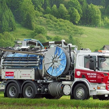 Ein großer rot-weißer LKW mit 'WILTON' auf der Seite steht auf einem grünen Feld mit einem Gebirgshintergrund.