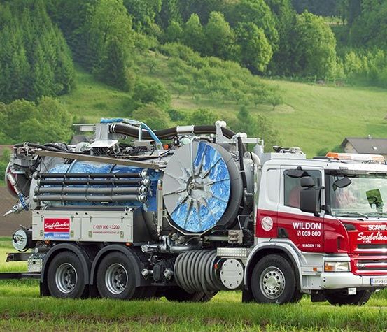Ein großer rot-weißer LKW mit 'WILTON' auf der Seite steht auf einem grünen Feld mit einem Gebirgshintergrund.