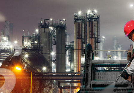 Ein Arbeiter in einem Chemiewerk bei Nacht, der eine Hochdruckreinigung verwendet und einen roten Helm sowie einen weißen Anzug trägt.