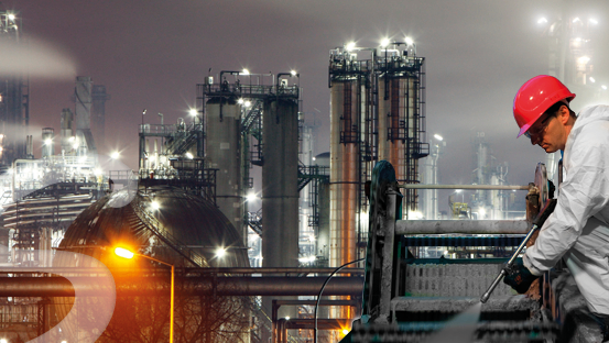 Ein Arbeiter in einem Chemiewerk bei Nacht, der eine Hochdruckreinigung verwendet und einen roten Helm sowie einen weißen Anzug trägt.