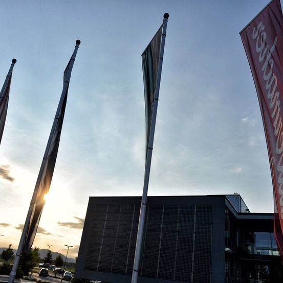 Drei Fahnen, darunter eine rote mit weißem Text, stehen vor einem Sonnenuntergangshimmel. Ein modernes Gebäude mit Solarpanelen an der Wand befindet sich im Hintergrund.