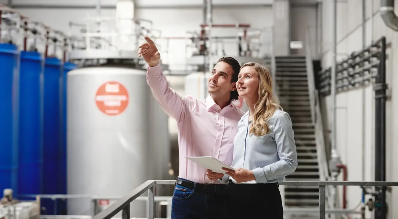 Ein Mann und eine Frau in einer Fabrikumgebung, der Mann zeigt auf Ausrüstung, während sie beide nach oben schauen. Der Mann trägt ein rosa Hemd und die Frau ein hellblaues Blusenshirt.