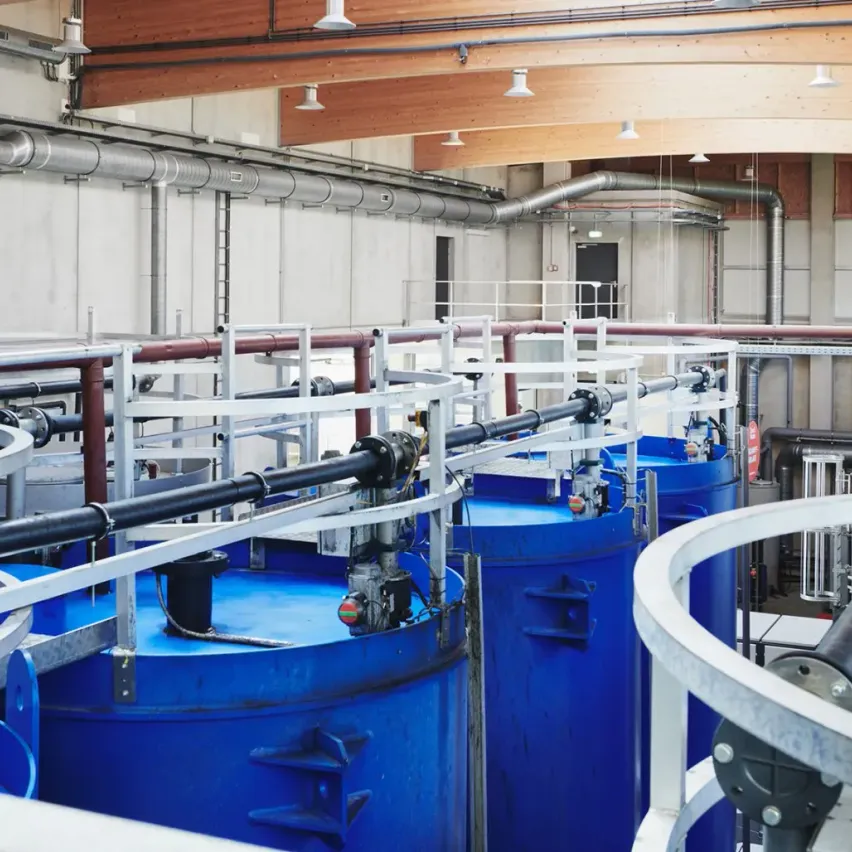 Industrieanlage mit großen blauen Tanks, die durch Rohre verbunden sind, unter einer Holzdecke mit hängenden Leuchten.