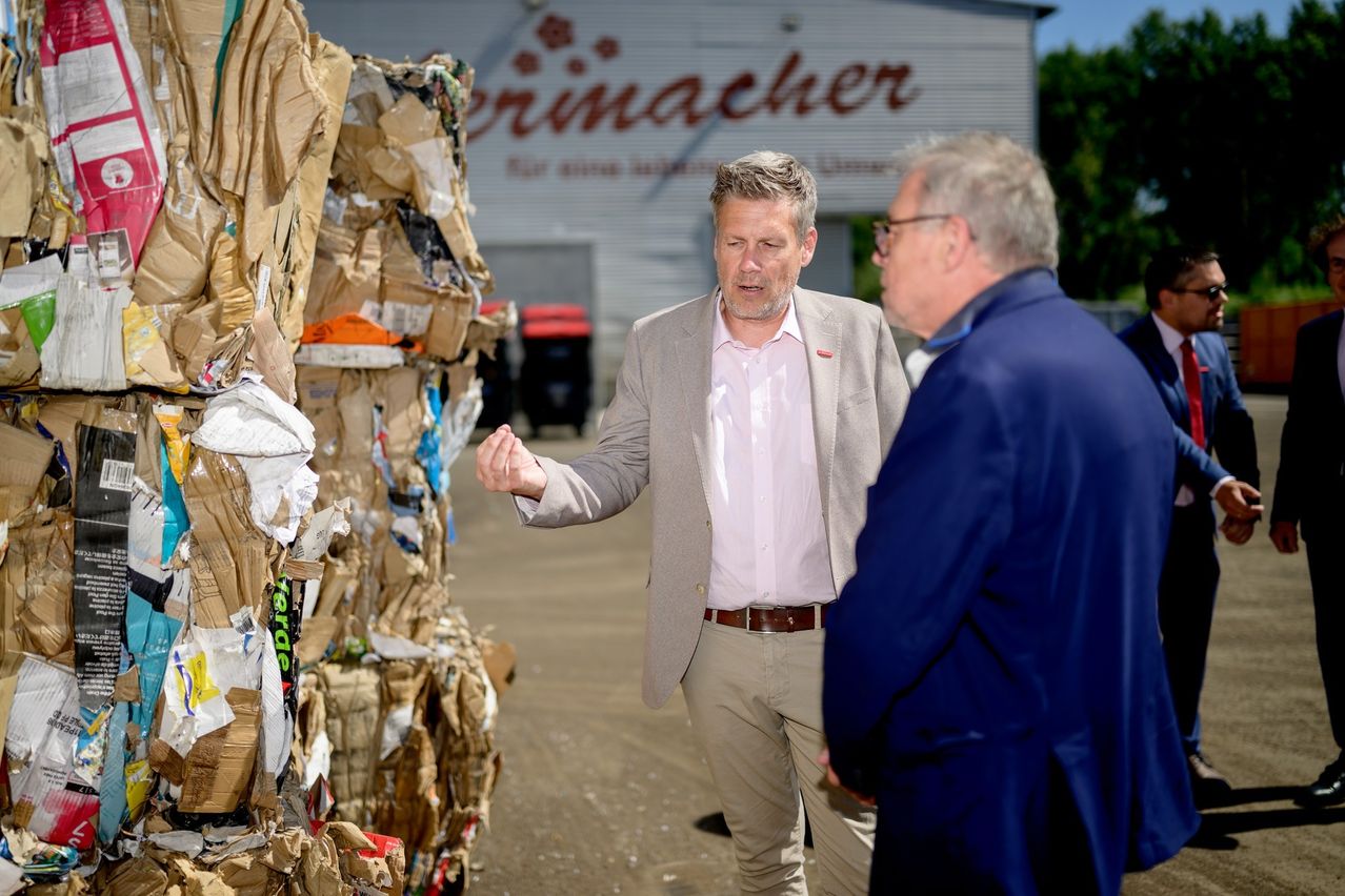 Zwei Männer in Anzügen diskutieren vor einem Gebäude mit der Aufschrift 'Ernamacher' und einem Haufen Pappkartons in der Nähe.