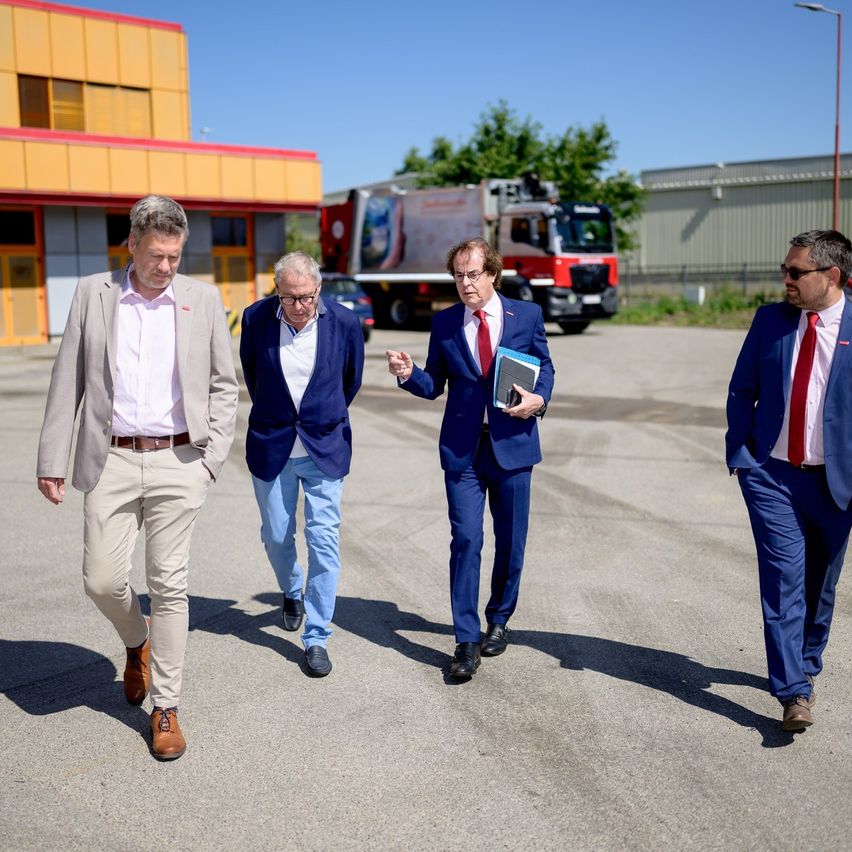 Fünf Männer in Anzügen gehen über einen Parkplatz, einer hält eine Mappe. Hinter ihnen ist ein Gebäude mit rotem Dach und ein Feuerwehrwagen zu sehen.