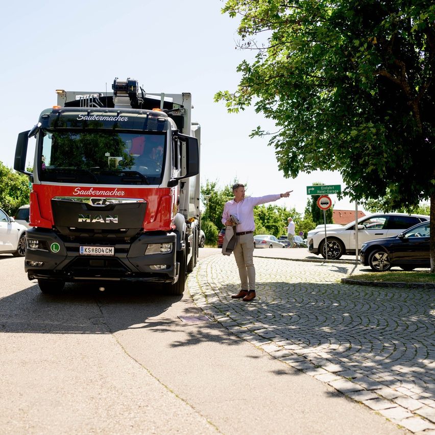 Ein Mann dirigiert einen großen roten MAN-Lkw mit einem Schild auf der Front. Mehrere Autos sind am Straßenrand geparkt.