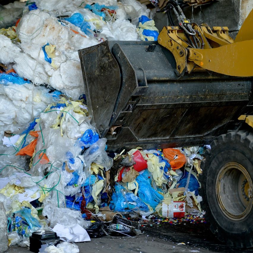 Ein gelber Maschinenwagen kippt einen großen Haufen Plastikabfälle auf den Boden. Der Abfall umfasst Plastiktüten, Flaschen und andere Gegenstände. Die Vorderseite des Fahrzeugs ist zur Kamera gerichtet.