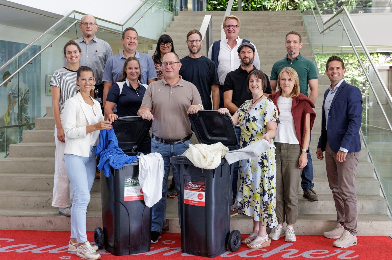 Eine Gruppe von Menschen steht auf einer Treppe neben Recycling-Behältern, wobei zwei Personen die Behälter öffnen. Sie sind lässig gekleidet, lächeln und scheinen für ein Foto zu posieren.