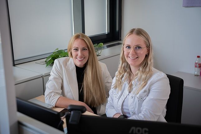Zwei Frauen sitzen an einem Schreibtisch in einem Büro, lächeln und schauen in die Kamera. Ein Computermonitor steht vor ihnen, und eine Coca-Cola-Flasche befindet sich auf einem nahegelegenen Schreibtisch.