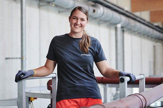 Eine Frau in einem blauen T-Shirt und orangefarbenen Hosen steht in einer Fabrik, lächelt und lehnt sich an ein Metallgeländer. Sie trägt Handschuhe und scheint für ein Foto zu posieren.
