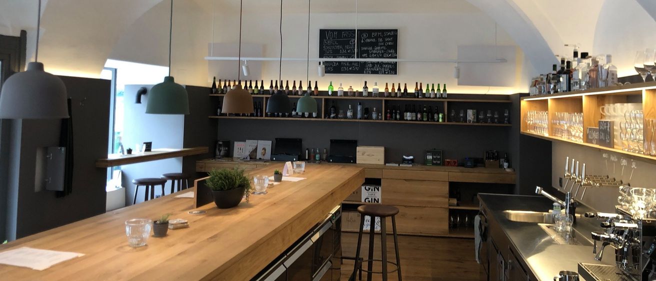 Eine Küche mit Holzschränken, einer Arbeitsplatte und Regalen voller Flaschen. Eine Tafel ist an der Wand montiert. Hängende Lampen und eine Topfpflanze schmücken den Raum.