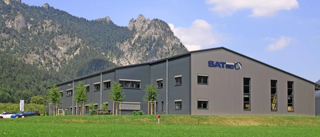 Ein modernes Gebäude mit dem SATec-Logo steht vor einer Berglandschaft unter einem blauen Himmel.