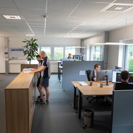 Ein Büroinnenraum mit vier Personen, drei stehend und einer am Schreibtisch sitzend, umgeben von Pflanzen, Computern und Bürogeräten, mit Fenstern, die einen Blick auf Bäume draußen bieten.