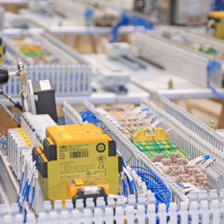 Ein industrielles Umfeld mit Reihen von elektronischen Komponenten, Kabeln und Steckverbindern. Ein gelbes Gehäuse mit Barcode ist im Vordergrund.