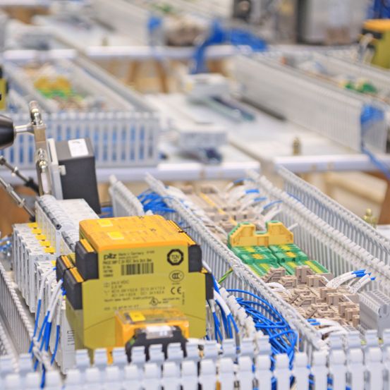 Ein industrielles Umfeld mit Reihen von elektronischen Komponenten, Kabeln und Steckverbindern. Ein gelbes Gehäuse mit Barcode ist im Vordergrund.