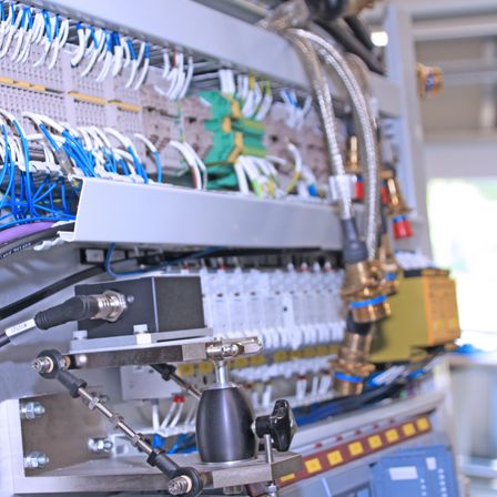 Eine Nahaufnahme einer industriellen Maschine mit verschiedenen Kabeln, Ventilen und Bedienfeldern. Die Maschine hat einen Metallrahmen und ist mit zahlreichen Kabeln und Rohren gefüllt.