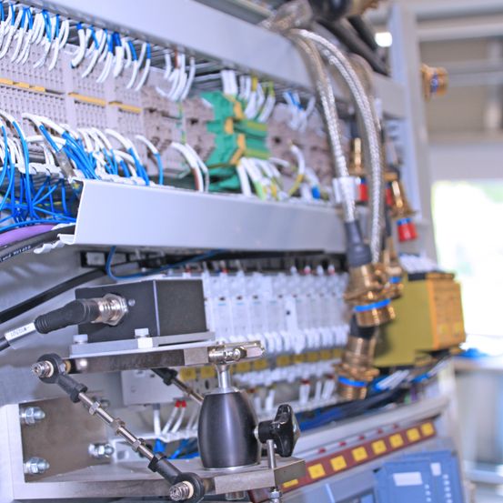 Eine Nahaufnahme einer industriellen Maschine mit verschiedenen Kabeln, Ventilen und Bedienfeldern. Die Maschine hat einen Metallrahmen und ist mit zahlreichen Kabeln und Rohren gefüllt.