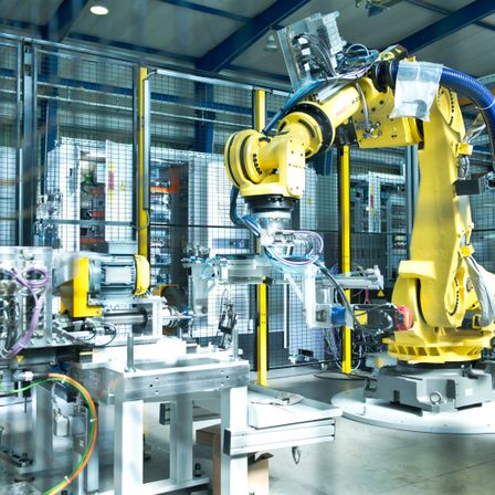 Ein industrieller Bereich mit gelben Robotern, die Teile zusammenbauen. Die Roboter sind mit Kabeln und Drähten verbunden. Der Bereich ist gut beleuchtet und von Metallzäunen umgeben.