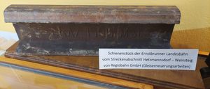 Ein verrostetes Metallstück mit Gravuren auf einem Holzschrank. Ein Etikett besagt: 'Schienenstück der Ernstbrunnen-Strecke von Regiobahn GmbH (Gleisereneuerung).'