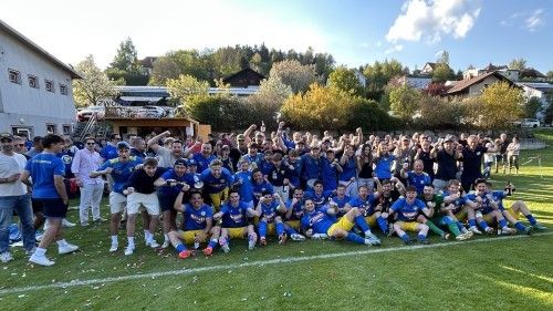 Ein Fußballteam in blauen und gelben Trikots feiert einen Sieg auf einem Rasenfeld vor Zuschauern und Bäumen im Hintergrund.