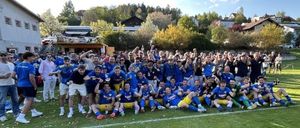 Ein Fußballteam in blauen und gelben Trikots feiert einen Sieg auf einem Rasenfeld vor Zuschauern und Bäumen im Hintergrund.