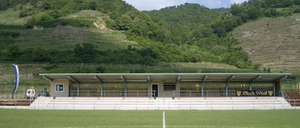 Ein leeres Fußballfeld mit einer weißen Linie, umgeben von einer Tribüne unter einem Dach, mit einem Berg im Hintergrund.