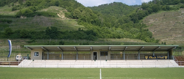 Ein leeres Fußballfeld mit einer weißen Linie, umgeben von einer Tribüne unter einem Dach, mit einem Berg im Hintergrund.