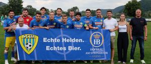 Eine Fußballmannschaft steht auf einem Feld mit einem blauen Banner, auf dem 'Echte Helden. Echte Bier.' steht. Die Teammitglieder tragen blaue Trikots mit dem Wort 'Schütz' und Nummern auf dem Rücken.