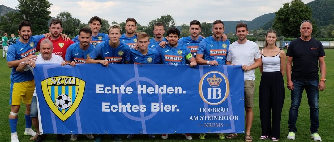 Eine Fußballmannschaft steht auf einem Feld mit einem blauen Banner, auf dem 'Echte Helden. Echte Bier.' steht. Die Teammitglieder tragen blaue Trikots mit dem Wort 'Schütz' und Nummern auf dem Rücken.
