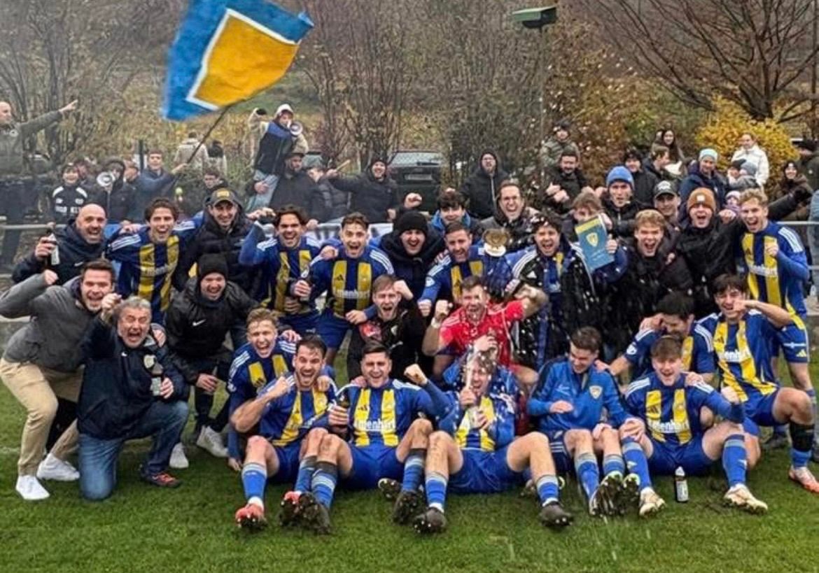 Eine Gruppe von Fußballspielern feiert einen Sieg auf einem Rasenfeld und hält einen Pokal. Hinter ihnen jubeln Fans und Spieler. Einige halten Fahnen und Getränke. Im Hintergrund befinden sich Bäume und ein Zaun.