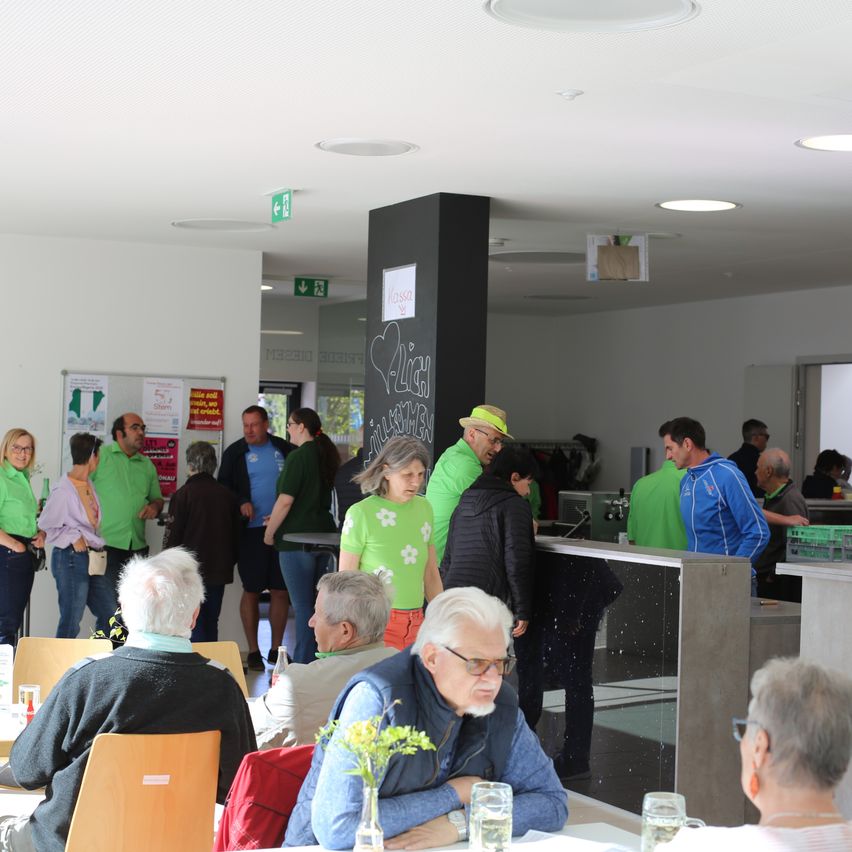 Eine Gruppe von Menschen in grünen Shirts und Hüten versammelt sich in einer Cafeteria. Einige sitzen an Tischen, während andere in der Nähe einer Theke stehen. Eine Frau mit einer gelben Mütze und Brille hält eine Vase mit gelben Blumen.