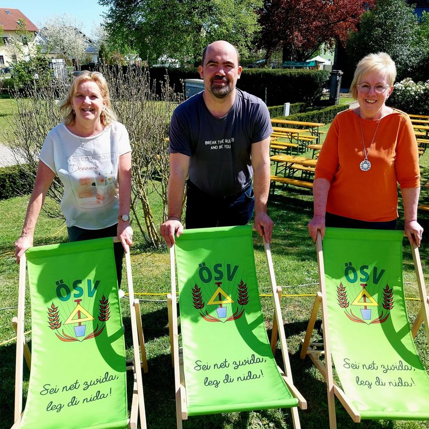 Drei Personen stehen in einem Außenbereich mit einem grünen Rasen und einigen Bäumen. Sie posieren für ein Foto und halten grüne Stühle. Die Stühle haben den Text Ã–SV und einige Wörter darauf geschrieben. Dahinter befinden sich einige gelbe Bänke und Bäume.