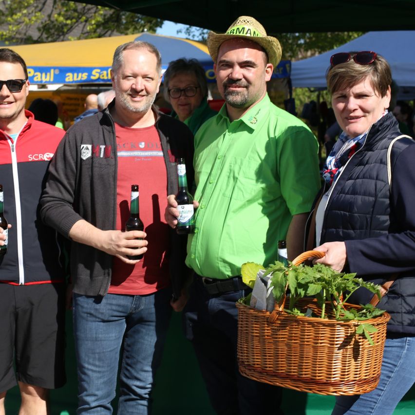 Vier Menschen stehen bei einem Outdoor-Event zusammen. Zwei Männer halten Flaschen, einer in grünem Shirt, einer in rotem Shirt. Eine Frau hält einen Korb mit Kräutern. Ein Mann im Hut lächelt.