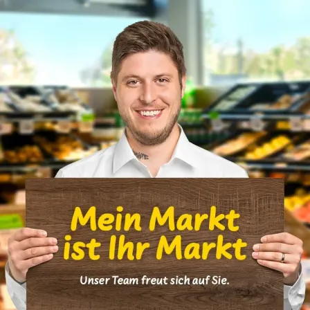 Ein lächelnder Mann in einem weißen Hemd hält ein hölzerne Schild, auf dem 'Mein Markt ist Ihr Markt' steht. Sein Team sieht glücklich aus. Der Hintergrund ist ein Supermarkt.