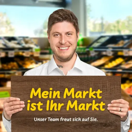 Ein lächelnder Mann in einem weißen Hemd hält ein hölzerne Schild, auf dem 'Mein Markt ist Ihr Markt' steht. Sein Team sieht glücklich aus. Der Hintergrund ist ein Supermarkt.