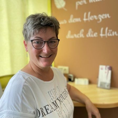 Eine Frau mit kurzen grauen Haaren und Brille lächelt und posiert für ein Foto. Sie trägt ein weißes T-Shirt mit schwarzem Text. Hinter ihr befindet sich eine Wand mit einem gelben Vorhang und einem Schreibtisch.