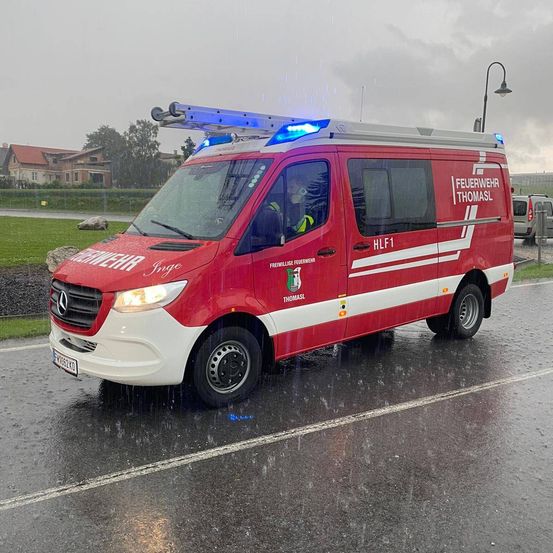 Ein roter Mercedes Benz Feuerwehrwagen fährt bei Regen auf einer nassen Straße. Das Fahrzeug hat oben ein blaues Licht und den Text 'FEUERWEHR THOMASL' an der Seite.