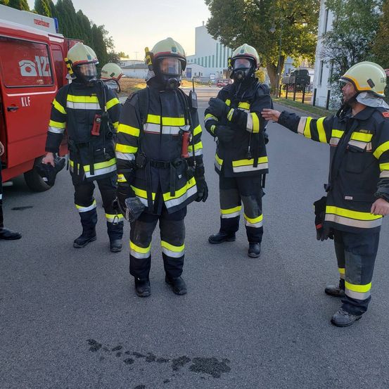 Feuerwehrleute in Schutzausrüstung stehen in einer Reihe auf der Straße. Sie unterhalten sich mit einem Mann vor ihnen. Ein roter Feuerwehrwagen ist hinter ihnen geparkt.