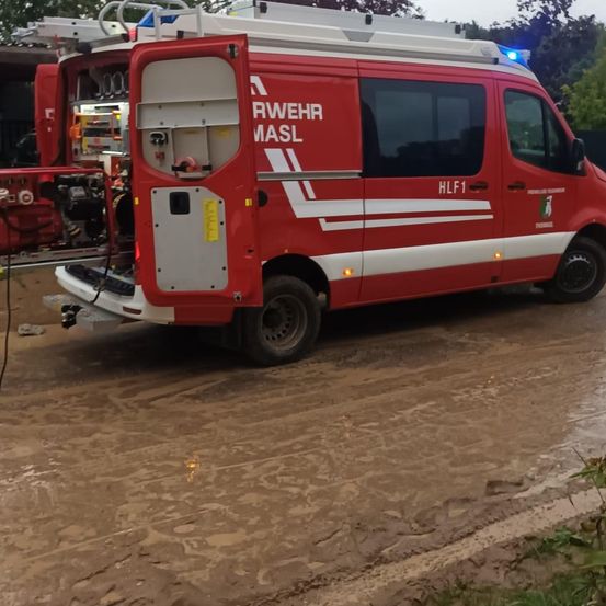 Ein roter Feuerwehrwagen mit der Aufschrift RWEHR MASIL und HLF1 an der Seite, der auf schlammigem Gelände mit offener Hecktür geparkt ist.