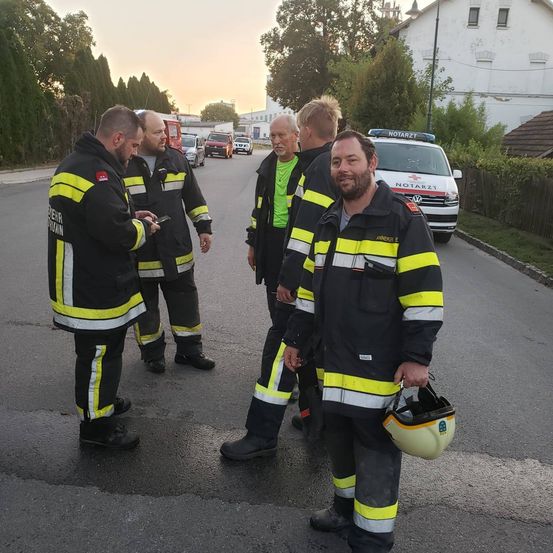 Fünf Feuerwehrleute stehen auf einer Straße, einer hält einen Helm. Einer schaut auf sein Handy. Hinter ihnen steht ein weißer Krankenwagen.