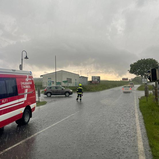 Ein Feuerwehrmann steht auf einer nassen Straße und regelt den Verkehr. Hinter ihm steht ein Feuerwehrwagen. Zwei Autos fahren die Straße entlang, während im Hintergrund ein Werbeschild zu sehen ist.