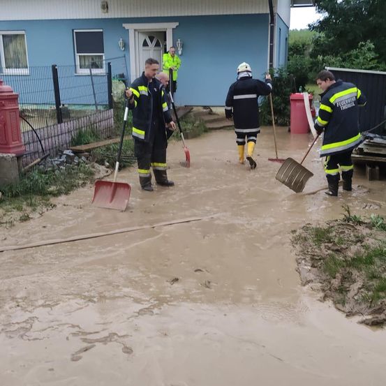 Mehrere Feuerwehrleute stehen in einem überfluteten Bereich, tragen Schutzanzüge, halten Schaufeln und Besen. Hinter ihnen steht ein blaues Haus mit einer Treppe, die zur Tür führt. Eine Person in einem gelben Schutzanzug steht auf der Treppe.