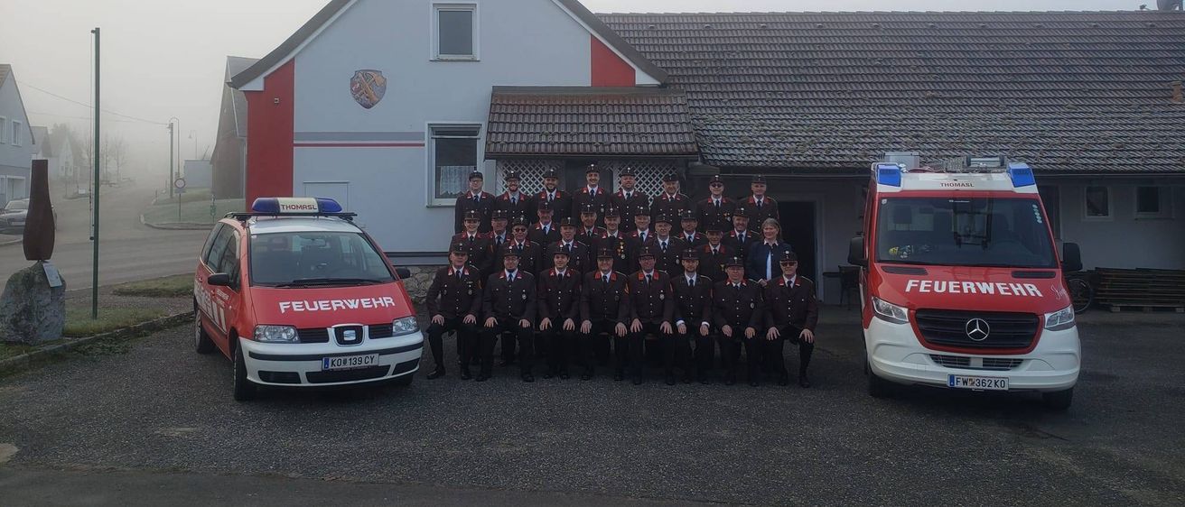 Eine Gruppe Feuerwehrleute in Uniform steht vor einem Feuerwehrauto und einem Polizeiwagen vor einem Haus mit einem Wappen an der Wand.
