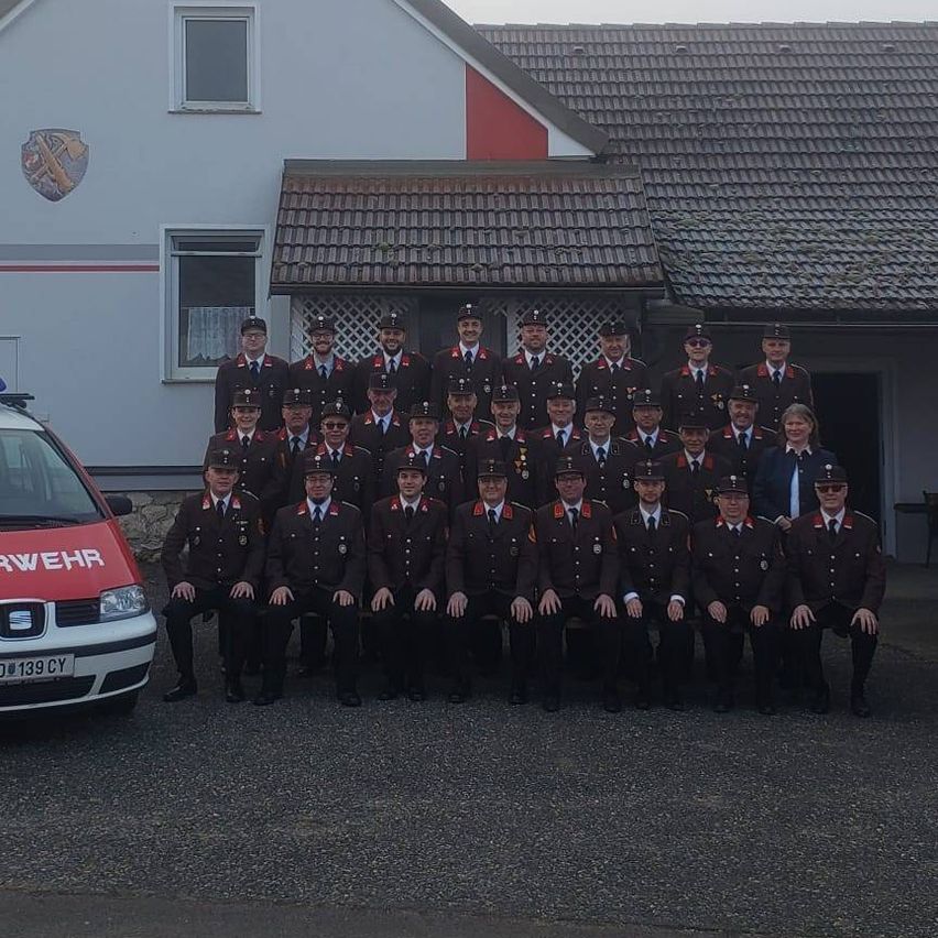 Eine Gruppe Feuerwehrleute in Uniform steht vor einem Feuerwehrauto und einem Polizeiwagen vor einem Haus mit einem Wappen an der Wand.