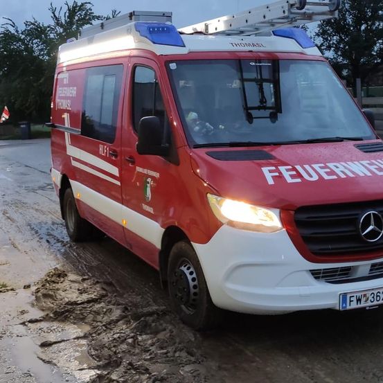 Ein roter Mercedes-Benz-Van, beschriftet mit 'Feuerwehr' und 'Thomasl', steht auf einer schlammigen Straße. Er hat eine Leiter auf dem Dach und seine Scheinwerfer sind eingeschaltet.