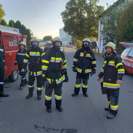 Fünf Feuerwehrleute in Schutzkleidung stehen draußen. Sie sind mit Helmen und Gasmasken ausgestattet. Zwei unterhalten sich, während andere sich umsehen. Ein roter Feuerwehrwagen ist hinter ihnen geparkt.