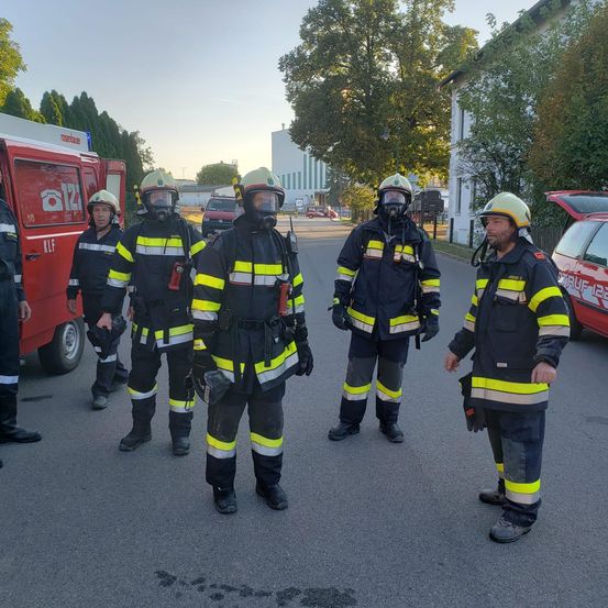 Fünf Feuerwehrleute in Schutzkleidung stehen draußen. Sie sind mit Helmen und Gasmasken ausgestattet. Zwei unterhalten sich, während andere sich umsehen. Ein roter Feuerwehrwagen ist hinter ihnen geparkt.