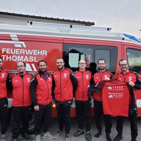 Sechs Männer in roten Jacken posieren vor einem Feuerwehrauto mit der Aufschrift 'Feuerwehr Thomasl'. Sie halten ein rotes T-Shirt mit den Texten 'STAD' und 'Feuerwehr Thomasl'.
