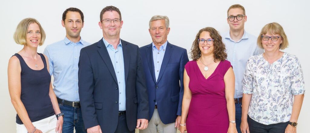 Eine Gruppe von Menschen in Geschäftskleidung steht eng beieinander und lächelt für ein Foto vor einer weißen Wand.